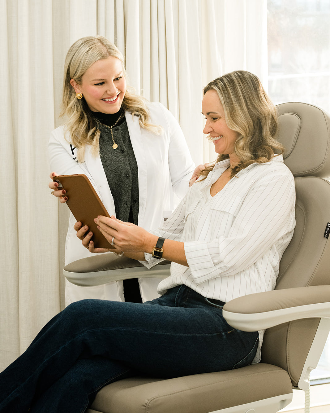 A woman sits in a beige dental or treatment chair while another woman in a white lab coat stands beside her holding a tablet. The scene is set in a bright, modern room with white curtains and a window.