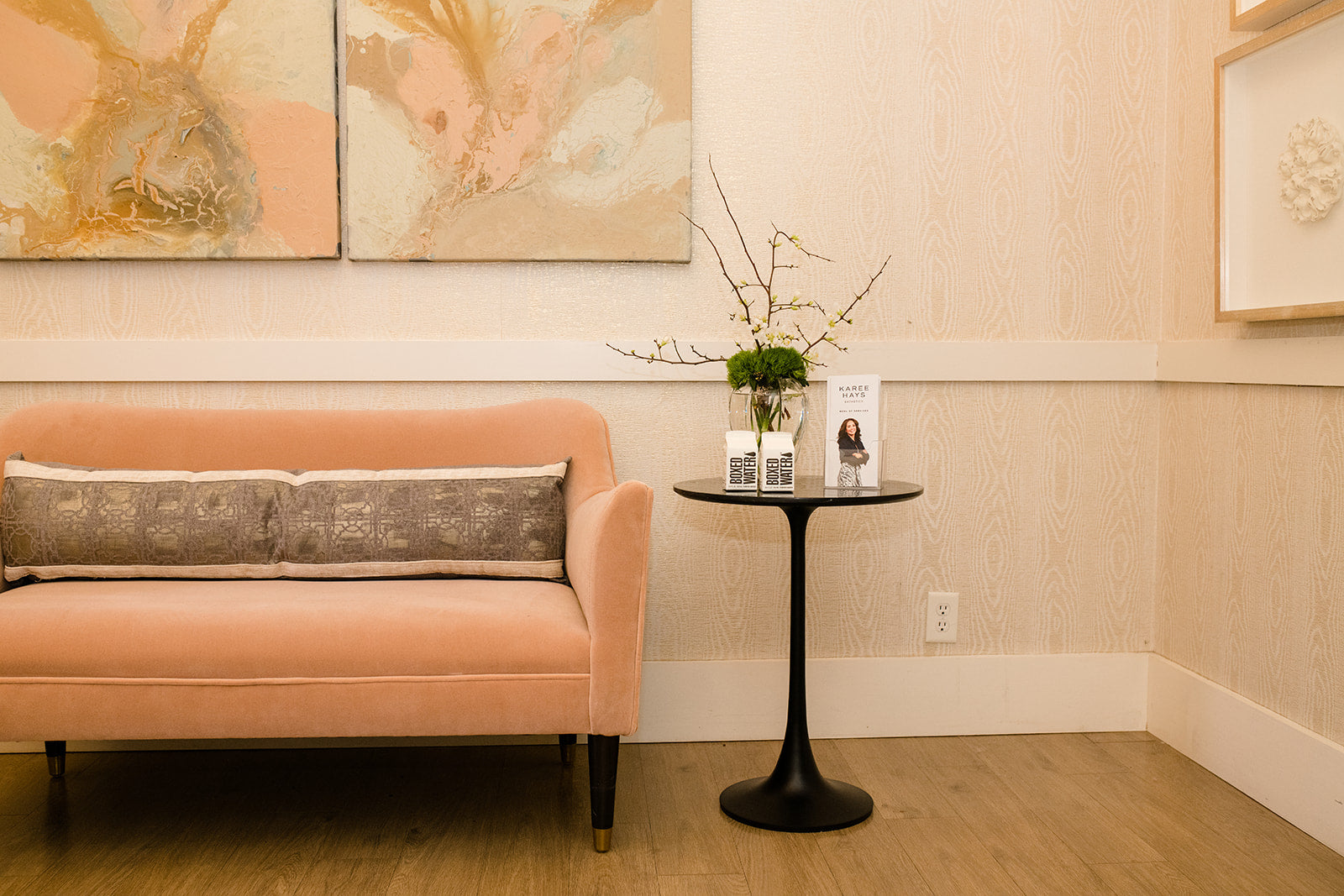 A peach-colored velvet sofa with two gray patterned throw pillows, positioned next to a small black round side table in a modern living room setting with beige wood-paneled walls and abstract wall art.